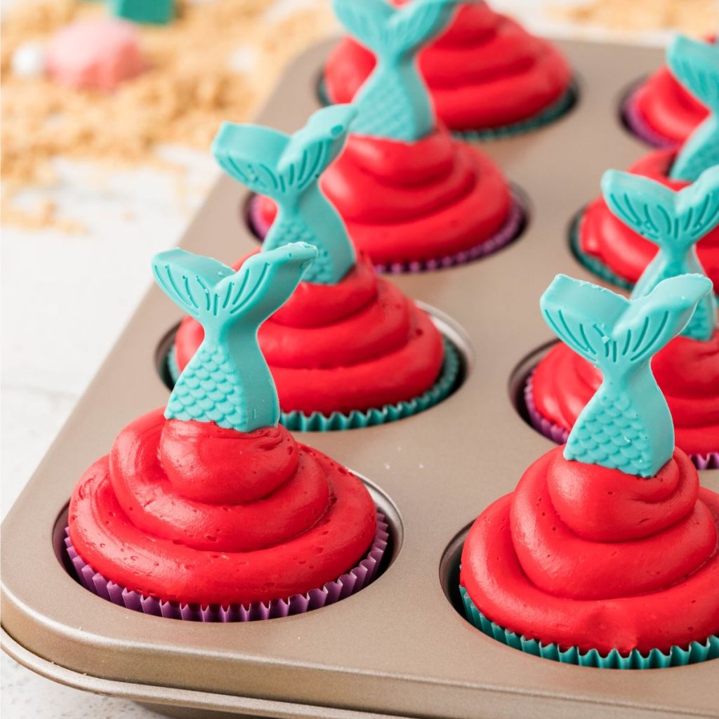Red frosted cupcakes with blue mermaid tail candy topper in muffin tin.