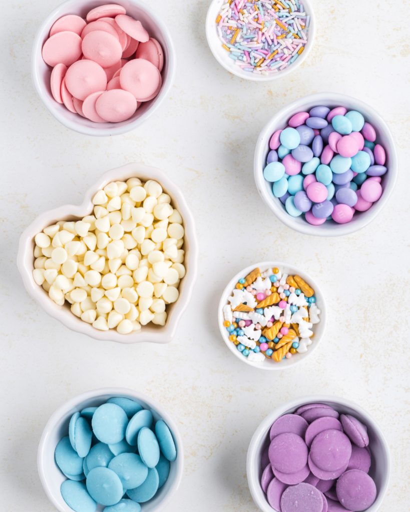 Bowls of pink, blue, and purple candy melts. Bowl of white chocolate chips and sprinkles. 