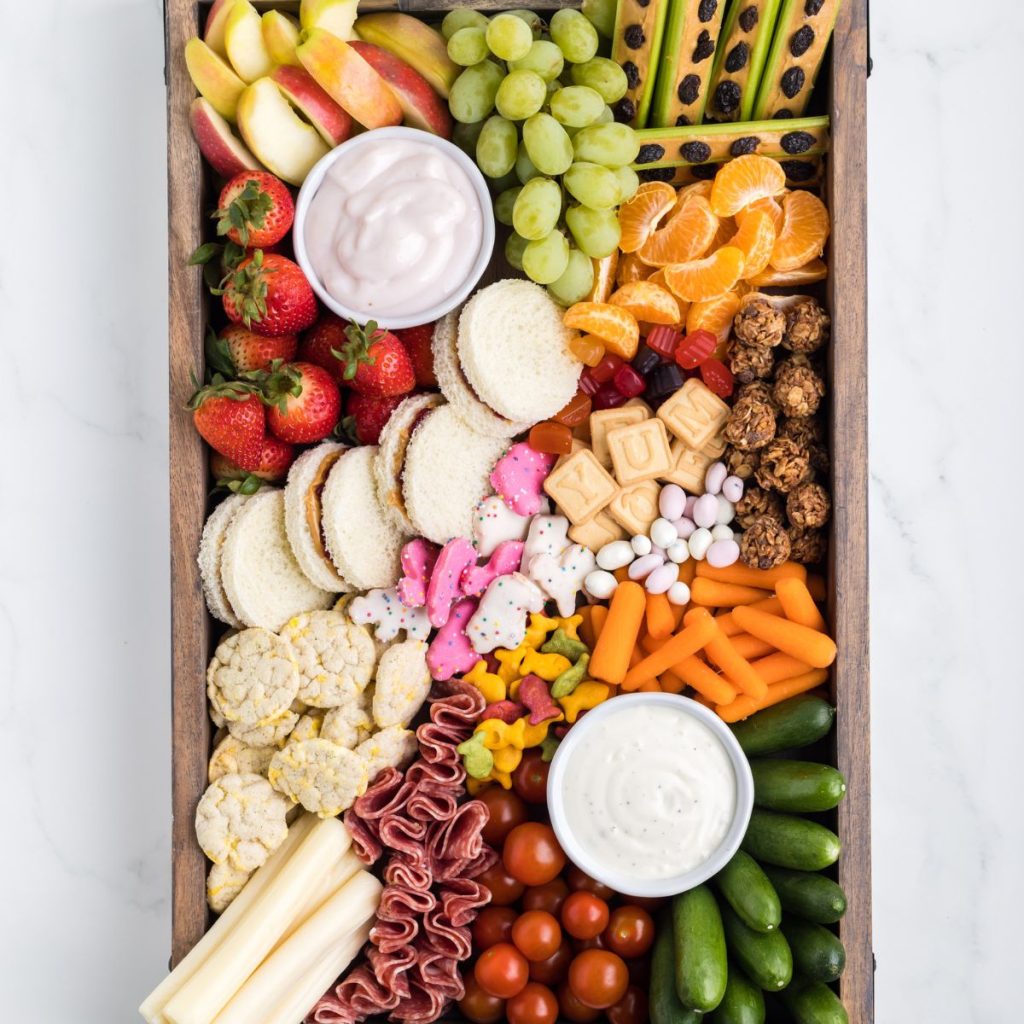 Board with sandwiches, fruit, vegetables, cookies, and dip. 