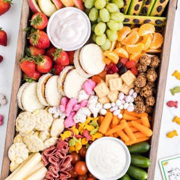 Snack board with grapes, vegetables, cheese, sandwiches, and dip.