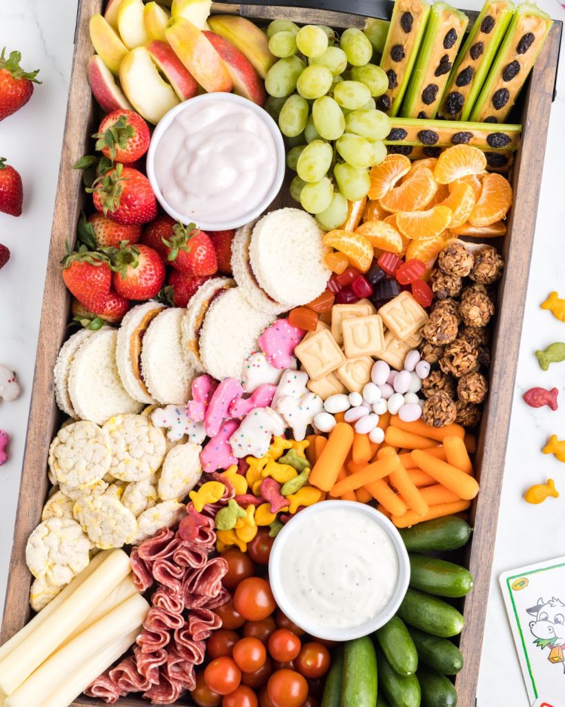Snack board with grapes, vegetables, cheese, sandwiches, and dip. 