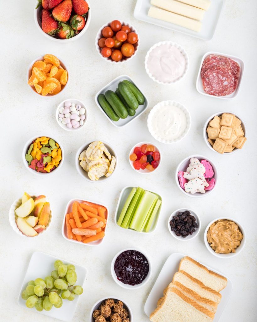 Bowl of fruit, vegetables, bread, deli meat, and cheese sticks. 