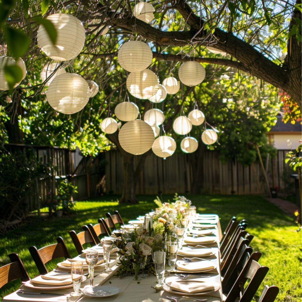Paper lanterns hanging in a tree over a table decorated with plates and flowers. 